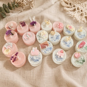 Op de afbeelding: Een verzameling decoratieve theelichtkaarsen in verschillende kleuren en ontwerpen. Sommige kaarsen zijn roze, andere wit, en sommige hebben bloemen of andere decoratieve elementen erop. De kaarsen zijn gerangschikt op een neutraal gekleurd oppervlak.