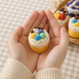 Op de afbeelding: Een kleine gele cake met witte glazuur en kleurrijke toppings, waaronder blauwe, groene en paarse bollen en stukjes chocolade. De cake wordt in komvormige handen gehouden, met andere soortgelijke cakes op de achtergrond.