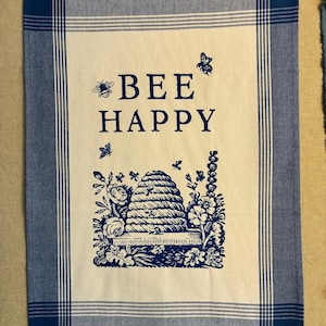 May include: A rectangular tea towel with a blue and white checked border. The centre features the words "BEE HAPPY" in navy blue, with a beehive and floral design. The towel has a light cream background.