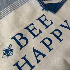 May include: White and blue kitchen towel with the words "BEE HAPPY" in dark blue block letters. A blue bee graphic is on the left side. The top of the towel has a blue and white striped border, and a blue plaid corner is folded over.