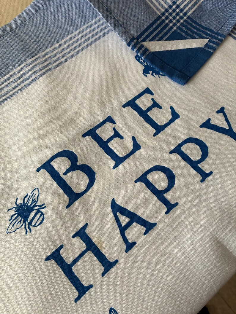 May include: White and blue kitchen towel with the words "BEE HAPPY" in dark blue block letters. A blue bee graphic is on the left side. The top of the towel has a blue and white striped border, and a blue plaid corner is folded over.