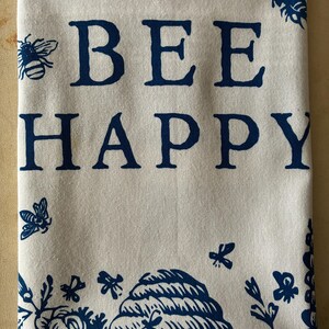 May include: A folded white tea towel featuring the phrase "BEE HAPPY" in dark blue capital letters. The towel is adorned with dark blue illustrations of bees, flowers, and a beehive. The design is repeated along the bottom and in the corners.