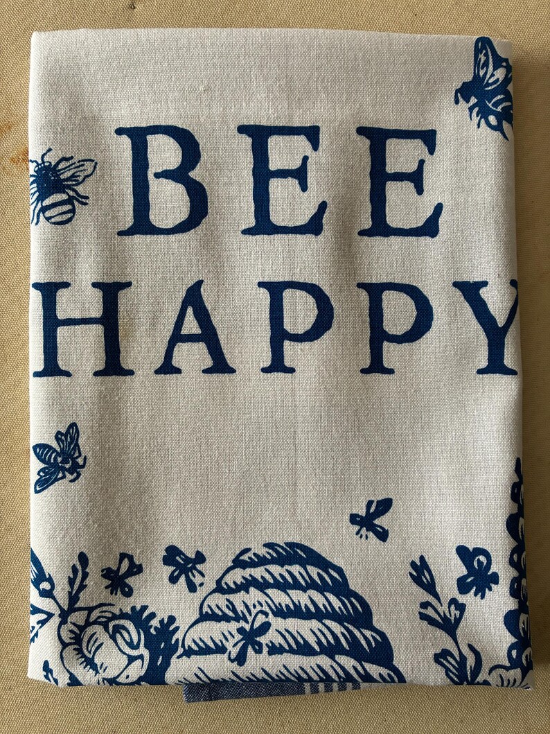 May include: A folded white tea towel featuring the phrase "BEE HAPPY" in dark blue capital letters. The towel is adorned with dark blue illustrations of bees, flowers, and a beehive. The design is repeated along the bottom and in the corners.