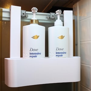 May include: A white over-the-door shower caddy holding two white Dove Intensive Repair shampoo bottles with gold pump dispensers. The caddy features a rectangular storage bin and two vertical support arms that hook over the top of a shower door frame.