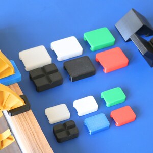 May include: A collection of small, square, multi-colored plastic corner protectors in white, black, green, red, and blue, alongside two larger black corner brackets and a yellow clamp securing a wooden board, arranged on a flat surface.