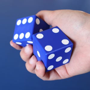 May include: Two blue dice with white dots are held in a hand. The dice are square-shaped and have a classic design. The background is a solid blue color. This image is suitable for games, gambling, or educational content.