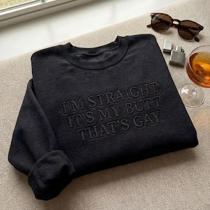 Peut inclure: Un sweat-shirt noir avec la phrase brodée "I'M STRAIGHT, IT'S MY BUTT THAT'S GAY". L'image présente également des lunettes de soleil, un briquet et un verre, posés sur une surface neutre.