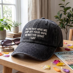 May include: A black baseball cap with white embroidered text that reads "PLS DO NOT GIVE ME A RIP OFF YOUR VAPE NO MATTER WHAT I SAY." The cap sits on a wooden table with sunglasses, a bracelet, and flower petals.