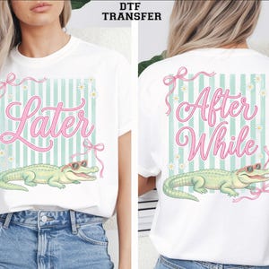 May include: A white t-shirt featuring a DTF transfer graphic of a light green alligator wearing red sunglasses. The design includes a light green and white vertical striped background, pink ribbons, small white daisies, and the text Later After While.