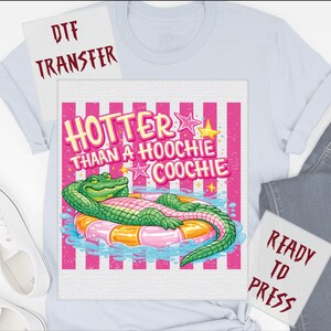 May include: Light blue t-shirt with a graphic of a green alligator relaxing on a pink and orange inflatable ring. The text "HOTTER THAN A HOOCHIE COOCHIE" is above the alligator in pink and yellow. DTF transfer.