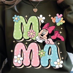 Puede incluir: Sudadera verde oliva con la palabra "MAMA" en letras grandes y coloridas con una fuente retro. El dise&ntilde;o incluye a Minnie Mouse, flores y estrellas. Las letras est&aacute;n delineadas en blanco y tienen un efecto cosido.