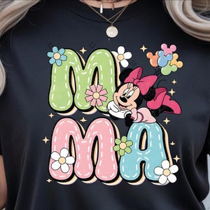 Puede incluir: Camiseta negra con la palabra "MAMA" en colores pastel y una fuente retro. Minnie Mouse se representa con un lazo rosa y globos. Acentos florales y estrellas rodean el texto, creando un dise&ntilde;o alegre.