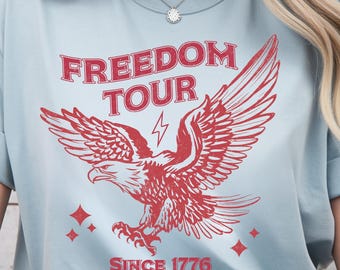 May include: Light blue t-shirt with a red graphic of an eagle with outstretched wings, a lightning bolt, and the words "FREEDOM TOUR" and "SINCE 1776". The design includes diamond shapes.