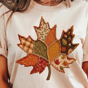 May include: A cream-colored t-shirt featuring a large, central maple leaf graphic. The leaf is composed of various fabric patches in autumn colors like orange, brown, green, and yellow, with visible frayed edges and decorative stitching.
