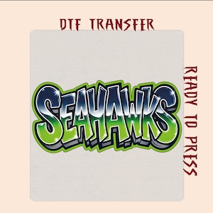 May include: A DTF transfer featuring the word SEAHAWKS in a stylized graffiti font with a gradient of dark blue to bright lime green. The letters have a white outline and a dark grey drop shadow. The text DTF TRANSFER and READY TO PRESS appears.