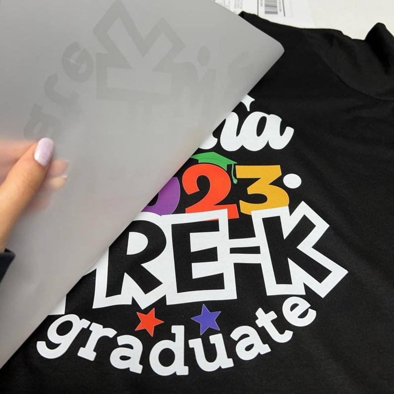 Puede incluir: Camiseta negra con un dise&ntilde;o colorido "Pre-K Graduate 2023". El dise&ntilde;o incluye un gorro de graduaci&oacute;n, estrellas y las palabras "Pre-K graduate" en negrita blanca. Se est&aacute; despegando una hoja de transferencia.