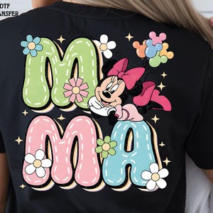 Puede incluir: Camiseta negra con la palabra "MAMA" en colores pastel, detalles florales y un dibujo de Minnie Mouse. Las letras tienen un contorno blanco cosido, y el dise&ntilde;o incluye estrellas.