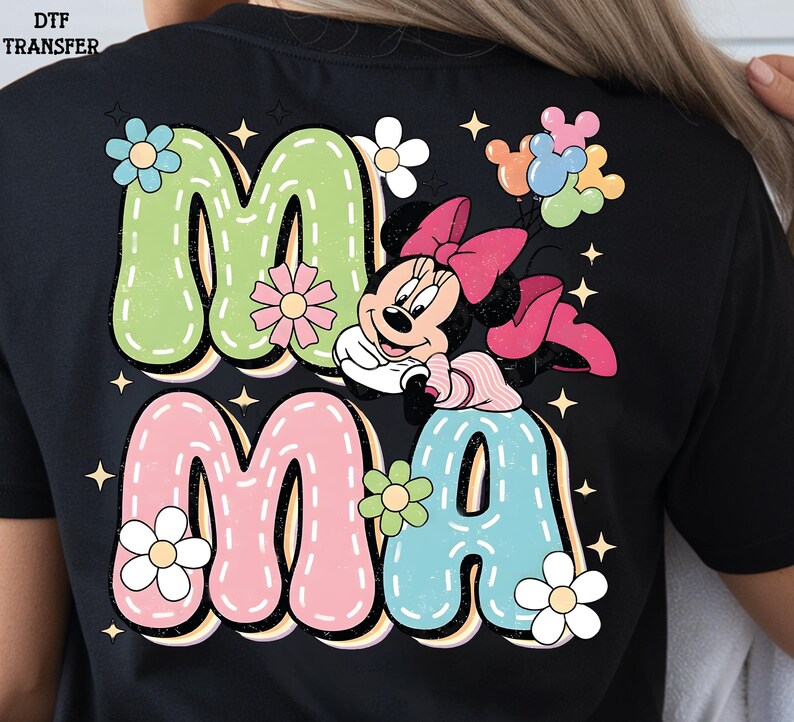 Puede incluir: Camiseta negra con la palabra "MAMA" en colores pastel, detalles florales y un dibujo de Minnie Mouse. Las letras tienen un contorno blanco cosido, y el dise&ntilde;o incluye estrellas.