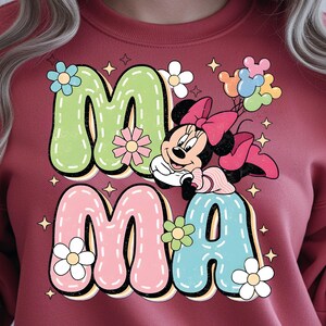 Puede incluir: Sudadera burdeos con la palabra "MAMA" en letras de burbujas. Las letras son verdes, rosas y azules, decoradas con flores y estrellas. Minnie Mouse aparece con un lazo rosa y globos.