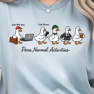 May include: A light blue t-shirt featuring a graphic of five white ducks. The first duck wears a red cap and holds a briefcase and laptop with the text data data data. The second duck holds a phone with the text Code Brown. The third wears a green beanie, the fourth a suit and tie, and the fifth holds a coffee cup. The text Para Normal Activities is printed below.