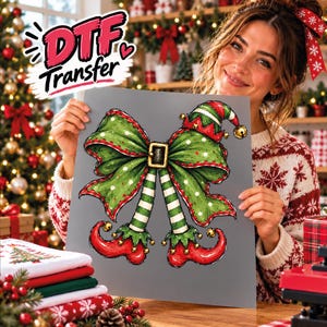 May include: A DTF transfer sheet featuring a graphic illustration of a large green bow with white polka dots, a black belt buckle, and two elf legs wearing striped stockings and curled red shoes with small gold bells attached to the tips.