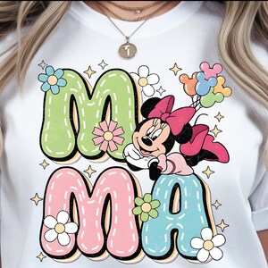 Puede incluir: Camiseta blanca con las palabras "MA MA" en letras coloridas. La "M" es verde, la "A" es rosa y la segunda "A" es azul. Minnie Mouse aparece con un lazo rosa y globos.