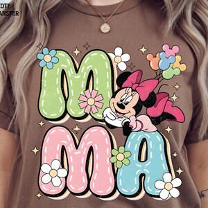 Puede incluir: Camiseta marr&oacute;n con la palabra "MAMA" en letras grandes y coloridas y un gr&aacute;fico de dibujos animados de Minnie Mouse. Las letras est&aacute;n delineadas en negro y decoradas con flores y estrellas. Minnie Mouse sostiene globos.