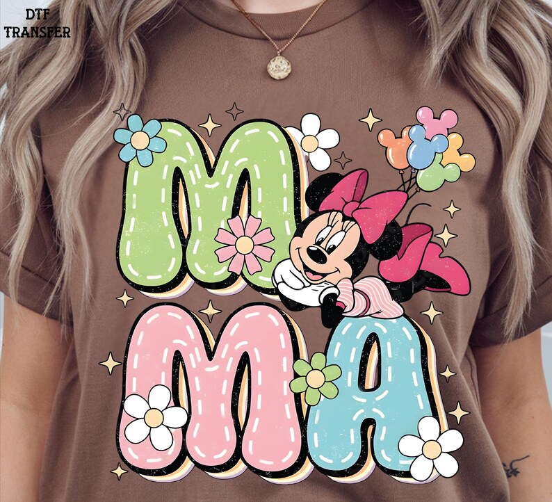Puede incluir: Camiseta marr&oacute;n con la palabra "MAMA" en letras grandes y coloridas y un gr&aacute;fico de dibujos animados de Minnie Mouse. Las letras est&aacute;n delineadas en negro y decoradas con flores y estrellas. Minnie Mouse sostiene globos.