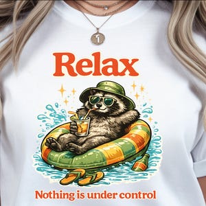May include: A white t-shirt featuring a graphic of a raccoon wearing a green bucket hat and sunglasses, floating in a green and orange striped inner tube in water. The raccoon holds a drink with a straw. Text reads Relax and Nothing is under control.