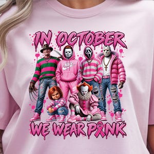 May include: A light pink t-shirt featuring a graphic of six horror movie characters dressed in pink clothing. The text IN OCTOBER WE WEAR PINK is printed in a stylized font. The design includes pink paint splatters and a pink awareness ribbon.