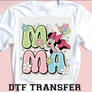 Puede incluir: Camiseta blanca con un gr&aacute;fico colorido. La palabra "MAMA" est&aacute; en letras de burbujas, decorada con flores y un personaje de dibujos animados sosteniendo globos. El texto "DTF TRANSFER" est&aacute; impreso en la parte inferior.