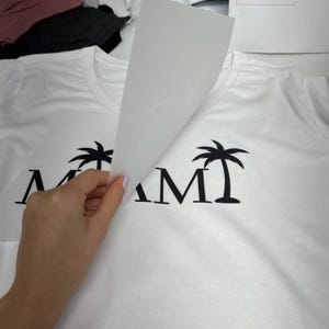 Puede incluir: Camiseta blanca con la palabra "MIAMI" en negro y un dise&ntilde;o de palmera negra. Una mano levanta una hoja de transferencia blanca, mostrando el dise&ntilde;o. La camiseta est&aacute; hecha de tela blanca suave.