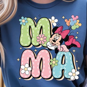 Puede incluir: Camiseta azul con la palabra "MAMA" en letras grandes y coloridas y un gr&aacute;fico de Minnie Mouse. Las letras est&aacute;n delineadas en negro y decoradas con flores y estrellas. Minnie Mouse sostiene globos.