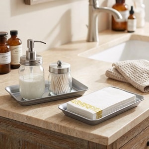 May include: A bathroom countertop arrangement featuring a metal tray with a soap dispenser, a jar of cotton swabs, and a tray of folded white paper napkins with a gold design. The soap dispenser is a mason jar with a silver pump.