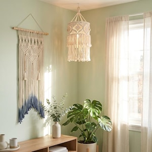 May include: A cream-colored macrame wall hanging with a blue dip-dyed bottom and a matching cream-colored macrame pendant light shade featuring intricate knotted patterns and long fringe, both suspended in a room with light green walls.