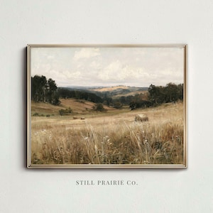 May include: A landscape painting in a thin gold frame depicting a golden wheat field with hay bales, rolling hills, and dark green trees under a cloudy sky. The text STILL PRAIRIE CO. is printed in black below the framed artwork on the wall.