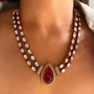 May include: A multi-strand necklace with a large, red, teardrop-shaped pendant. The necklace features white and red gemstones, with gold-coloured metal detailing. The jewellery is worn around the neck.