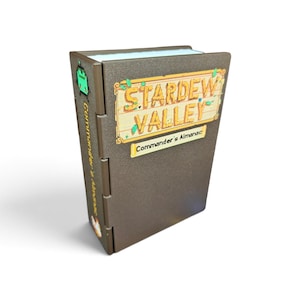 May include: A brown, rectangular, hinged box resembling a book with the text Stardew Valley and Commander's Almanac on the front cover. The spine features a small green character graphic and the text Commander's Almanac in yellow lettering.