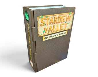 May include: A brown, rectangular, hinged box resembling a book with the text Stardew Valley and Commander's Almanac on the front cover. The spine features a small green character graphic and the text Commander's Almanac in yellow lettering.