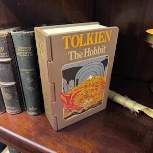 Può includere: Una scatola rettangolare a forma di libro di colore marrone con la scritta "TOLKIEN The Hobbit" stampata in giallo sul davanti. Il pannello frontale presenta un'illustrazione colorata di un drago rosso su un mucchio di monete d'oro.
