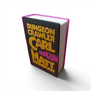 May include: A hardcover book titled Dungeon Crawler Carl by Matt Dinniman. The cover is black with gold embossed lettering and a pink jagged graphic element. The spine and edges of the book are a vibrant pink, contrasting with the dark cover.