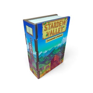 May include: A rectangular box designed to resemble a book, featuring the Stardew Valley Commander's Almanac title on the front cover. The exterior displays a colorful landscape illustration with mountains, a red barn, and blue sky.