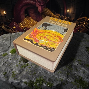 Könnte beinhalten: Ein gebundenes Buch mit dem Titel The Hobbit von Tolkien mit beigefarbenem Einband. Das Cover zeigt eine rechteckige Illustration eines roten Drachen, der auf einem Haufen Goldmünzen liegt. Das Buch hat weiße Seiten und liegt schräg.