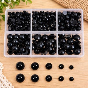 May include: A clear plastic storage container with six compartments filled with various sizes of round, glossy black beads. Below the container, ten individual black beads are arranged in two rows, showcasing a range of different diameters.