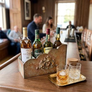 May include: A rectangular, weathered copper-toned metal caddy with an ornate central crest, holding four glass bottles of whiskey including The Macallan 18, Glenfiddich, and Jameson. Beside the caddy sits a small gold tray with two crystal glasses.