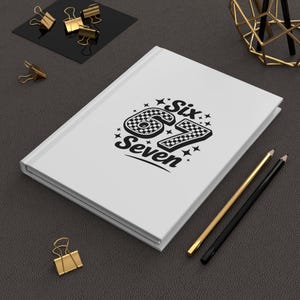 May include: A white hardcover notebook featuring a black graphic design on the front cover. The design displays the text Six 67 Seven with the numbers 67 filled with a black and white checkered pattern, surrounded by small star shapes.