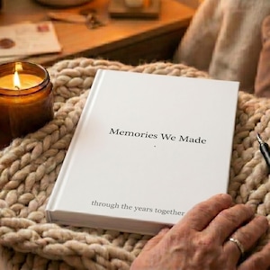 May include: A white hardback book with the black printed text Memories We Made and through the years together on the front cover. The book is positioned on a thick beige knit fabric next to a lit candle in a glass jar and a black pen.