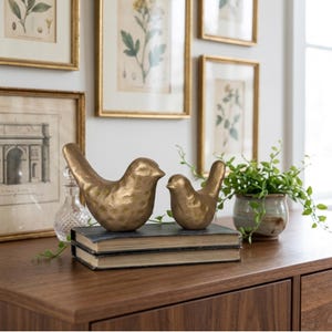 May include: Two gold bird ornaments, a large and a small one, rest on old books. The birds are on a dark wooden surface, with framed botanical prints in the background. A small potted plant is also visible.