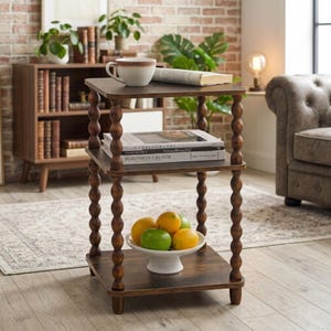 May include: A three-tiered, dark brown side table with turned legs. The top shelf holds a white and brown mug and a book. The middle shelf has books, and the bottom shelf displays a white pedestal bowl with oranges and a green apple.