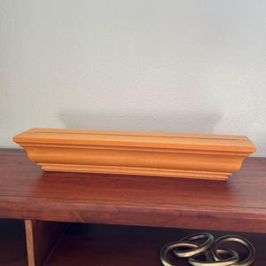 Puede incluir: Un soporte de exhibición rectangular de madera de color naranja claro con una ranura horizontal en la superficie superior. El soporte presenta un diseño de base moldeada y escalonada con bordes lisos y limpios, y un acabado mate uniforme.